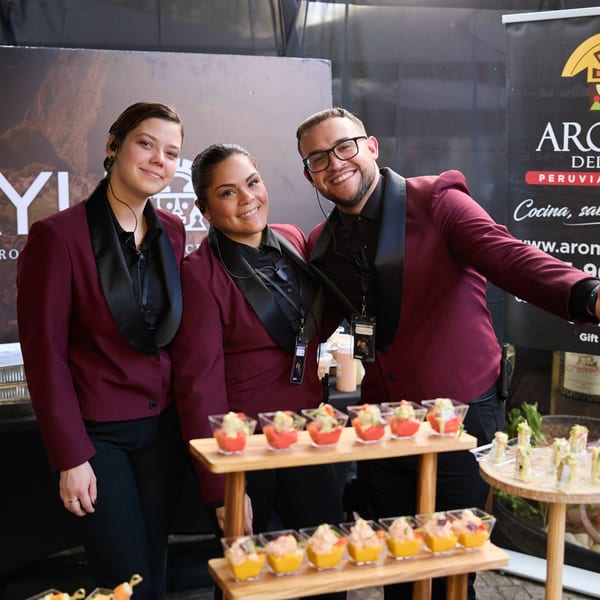 Three smiling servers in maroon blazers stand behind a table of small appetizers.