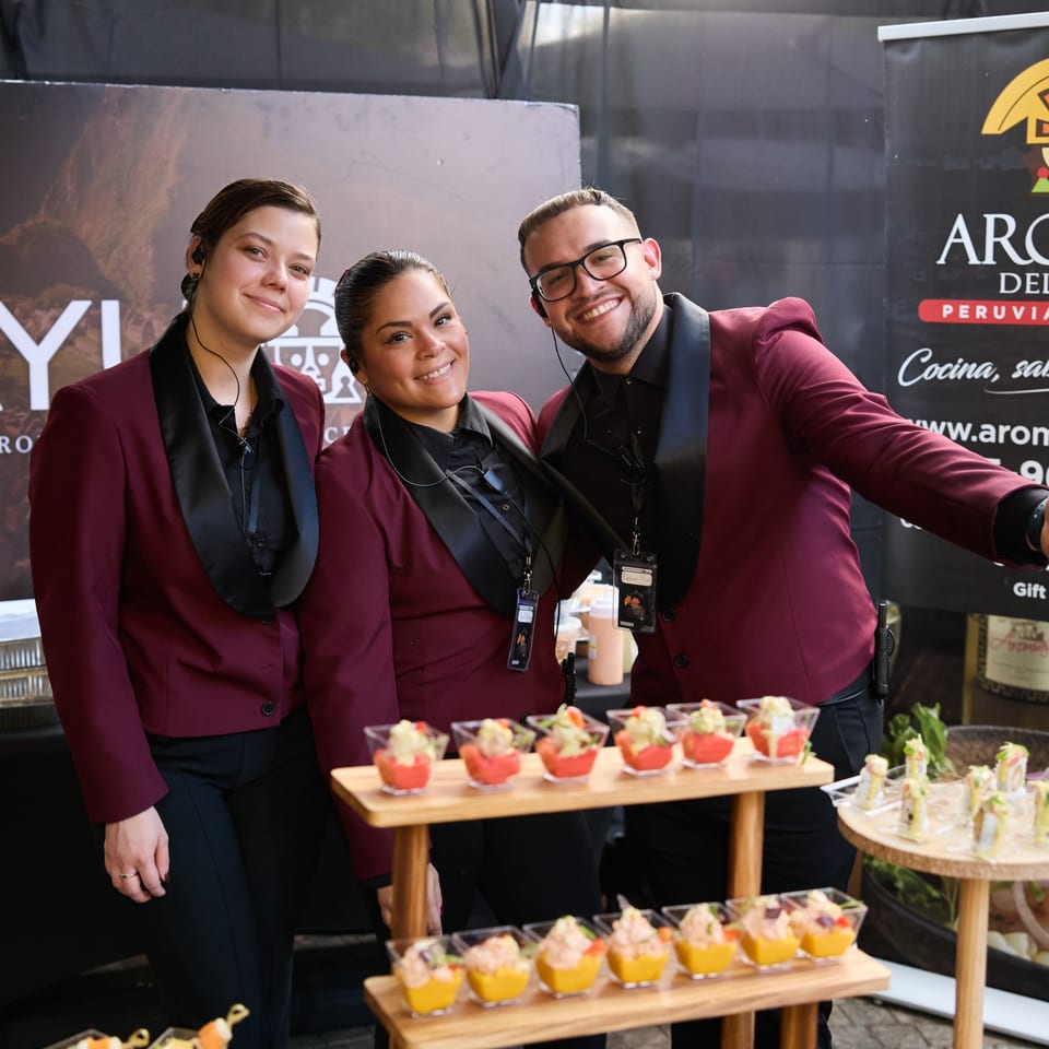 Three smiling servers in maroon blazers stand behind a table of small appetizers.