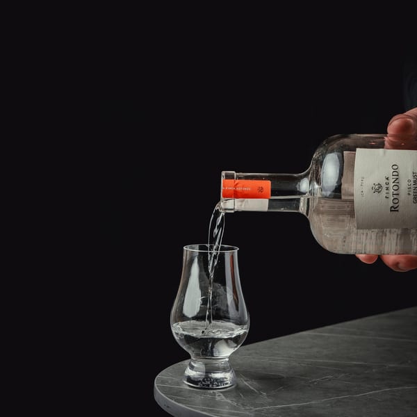 A hand pours clear liquid from a Finca Rotondo Pisco bottle into a glass on a dark table.