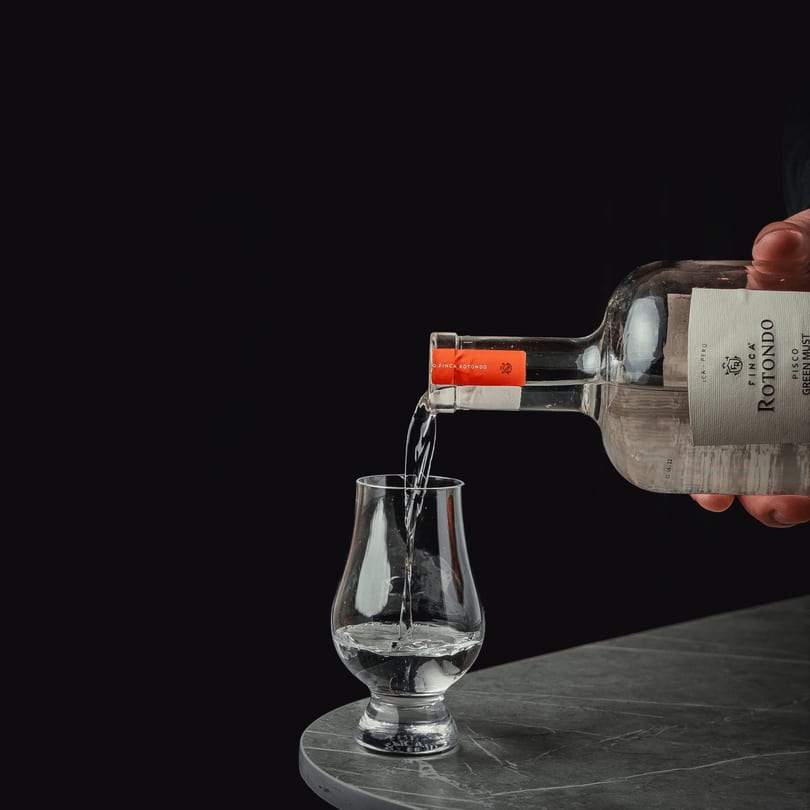 A hand pours clear liquid from a Finca Rotondo Pisco bottle into a glass on a dark table.