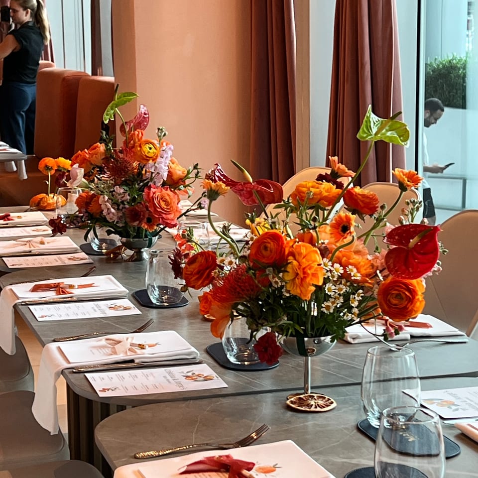 A long dining table set with white plates, menus, glasses, and vibrant orange floral centerpieces.