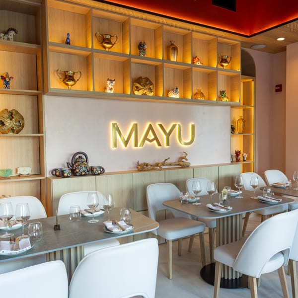 The Mayu restaurant interior features a glowing "MAYU" sign, light wood shelving with decor, and set tables.