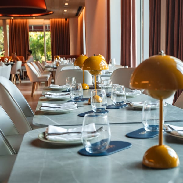 A modern restaurant dining room with set tables, yellow lamps, and large windows with outdoor views.