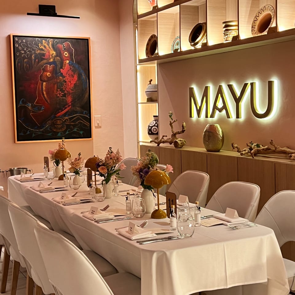 A long dining table set for guests with floral arrangements and a glowing "MAYU" sign.