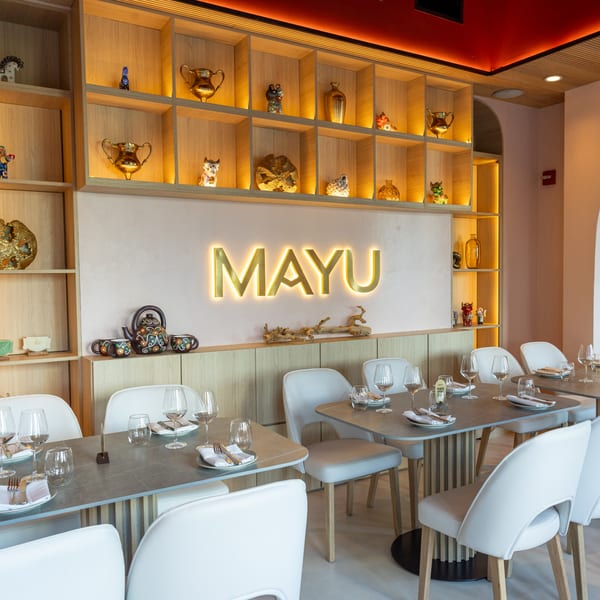 Modern restaurant interior with a glowing "MAYU" sign, wooden shelves with decor, and set dining tables.