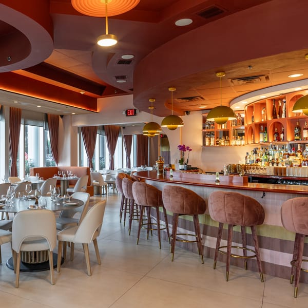 A stylish restaurant interior with a curved bar, pink velvet bar stools, and dining tables.