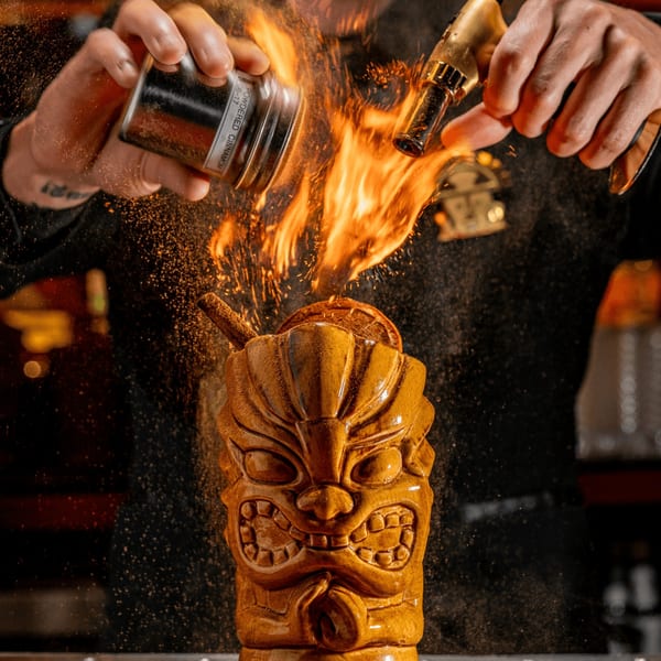 Bartender flaming a tiki cocktail with cinnamon powder and a torch, creating sparks and fire.