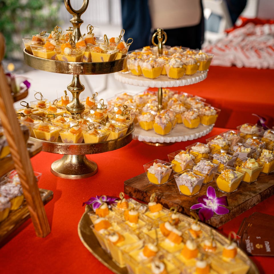 A vibrant dessert spread with mango and taro puddings in small cups on tiered stands and wooden boards.