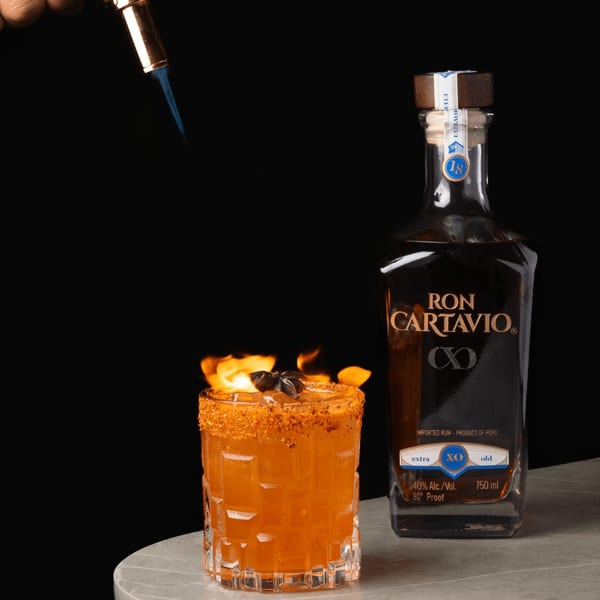 A flaming cocktail with a spiced rim and star anise, next to a bottle of Ron Cartavio rum.