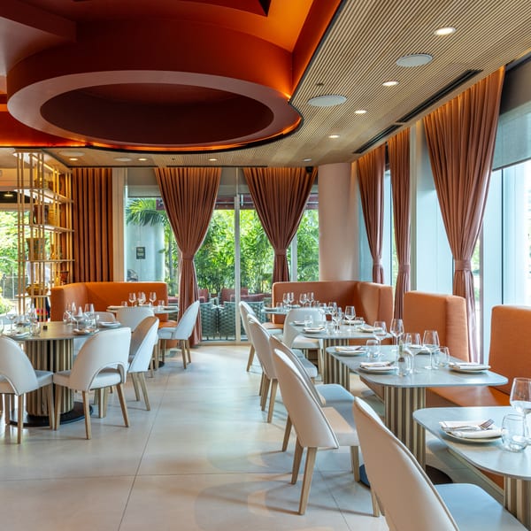 Modern restaurant dining room with orange accents, set tables, and large windows overlooking lush greenery.
