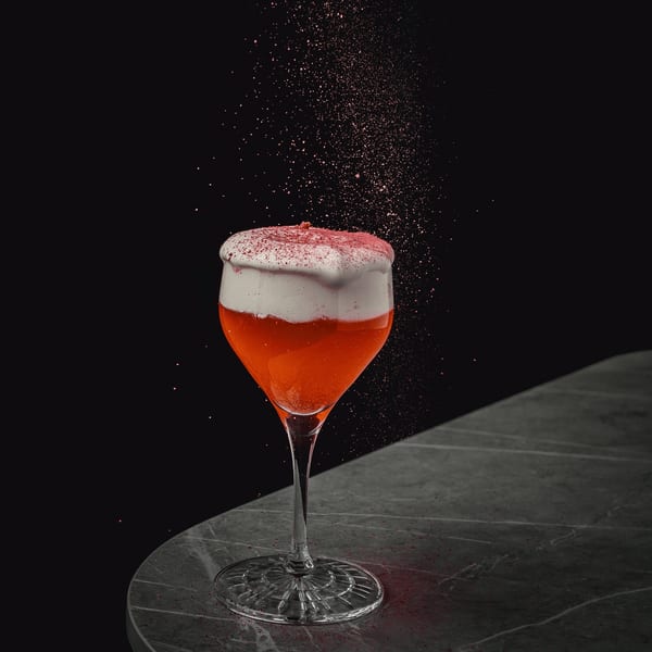Red cocktail with white foam and red sprinkles falling, on a dark marble table.