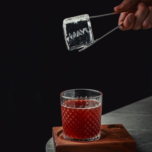Hand using tongs to place a large ice cube with "MAYU" etched into it into a red drink.