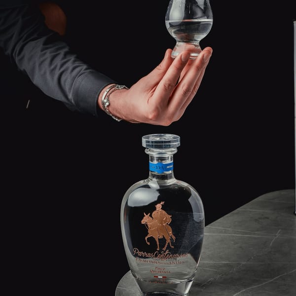A hand holding a glass of clear liquid above a bottle of Pisco Quebranta on a dark surface.